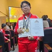 Albert muestra un diploma y varias medallas, vestido con un conjunto deportivo rojo.