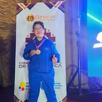 Albert muestra una medalla frente a un panel de Binaria relacionado con un concurso de matemática.